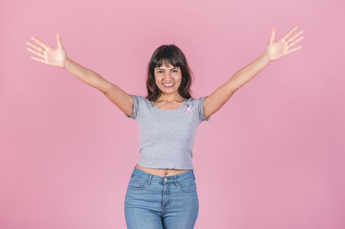 Cheerful woman with open arms wearing a breast cancer pink ribbon over pink background.  Victory to breast cancer concept.