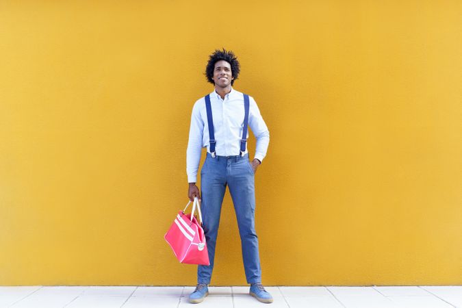 Man standing tall with curly hair wearing shirt and suspenders walking next to yellow wall