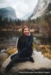Smiling woman in dark bubble jacket sitting on rock formation near lake 4N3Mgb