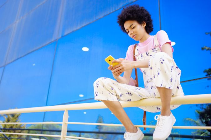 Woman sitting on handrails in front of reflective building checking phone
