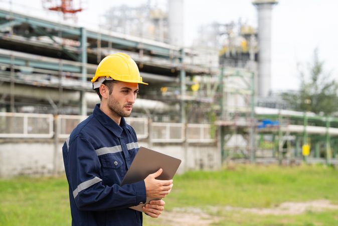 Refinery oil engineer worker in protection hardhat with laptop inspecting at petrochemical factory.