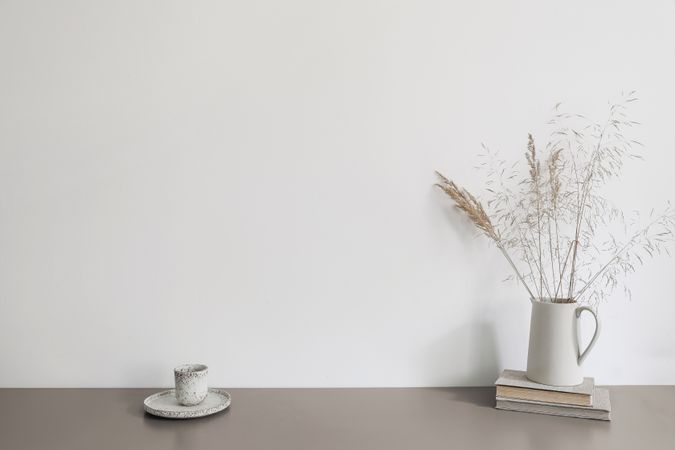 Autumn breakfast still life scene. Ceramic jug with dry grass and old books. Cup of coffee on beige table.