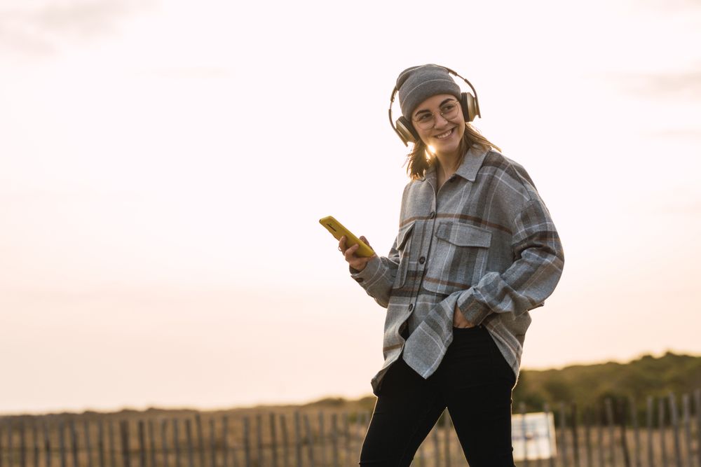 Happy young woman using smartphone and listening to music in headphones in countryside
