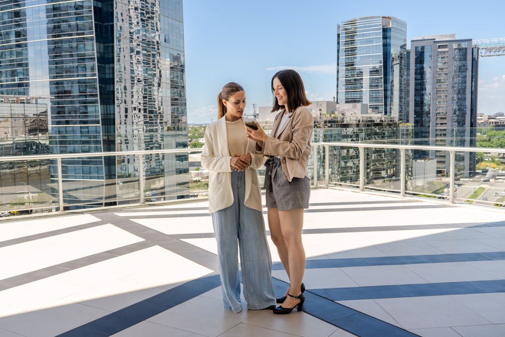 Two businesswomen standing on a terrace, one of them holding a cell phone and showing an email to her colleague. The scene is set in a city with tall buildings in the background.