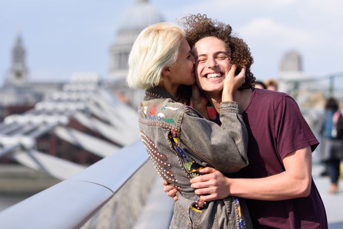 Blonde female kissing her smiling boyfriend on bridge in London