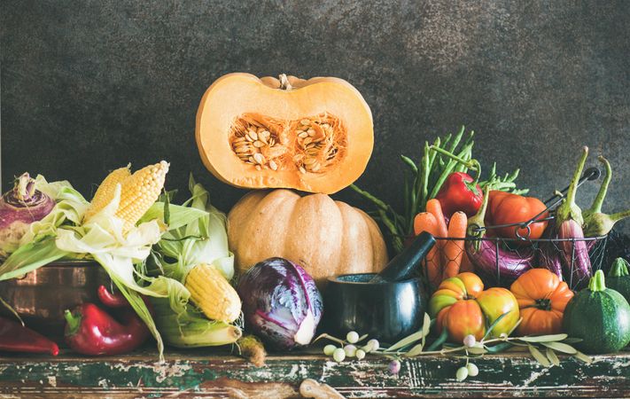 Fresh produce against dark backdrop with central halved squash, corn, cabbage, heirloom tomatoes