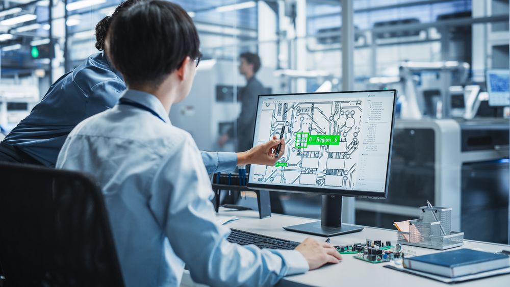 Hispanic Female Manager And Asian Male Engineer Using Desktop Computer With Microchip Blueprints at Automated Factory With Robotic Arms.