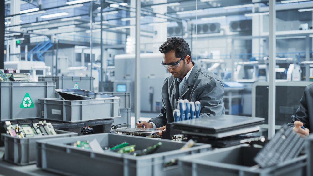 Indian Male Worker Disassembling Old Laptops To Recycle Electronic Components For Microchip Production At Automated Electronics Factory With Robotic Arms.