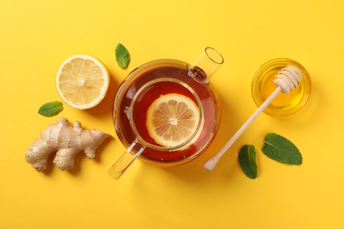 Flat lay. Teapot, lemon, ginger, mint, honey and dipper on yellow background, copy space