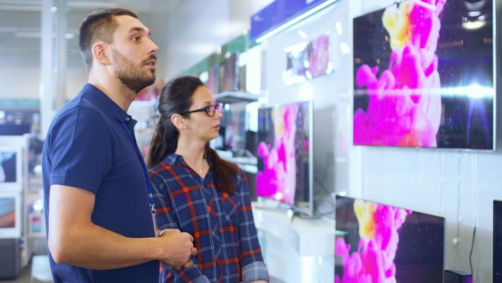 In the Electronics Store Professional Consultant Provides Expert Advice to a Young Woman Who Looks for TV Models