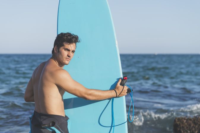 Male surfer holding blue board standing in front of the sea and looking back at camera