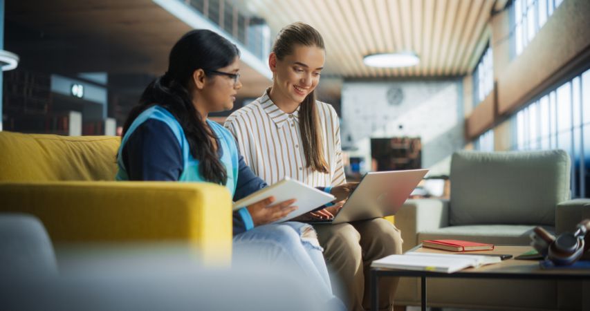 Two Diverse Friends and Classmates Working on a Collaborative Team Project in an Academic Library. Young Indian and White Women Using Laptop Computer for Online Learning and Virtual Lectures