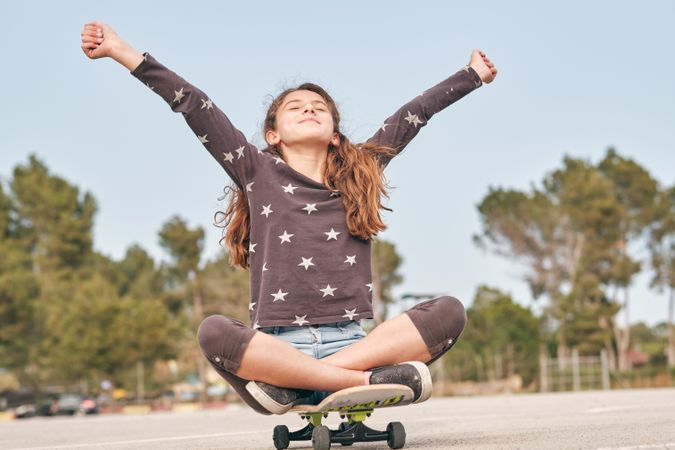 Excited girl sitting with outstretched arms on skateboard