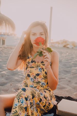 Woman sitting and sniffing rose at beach looking at camera