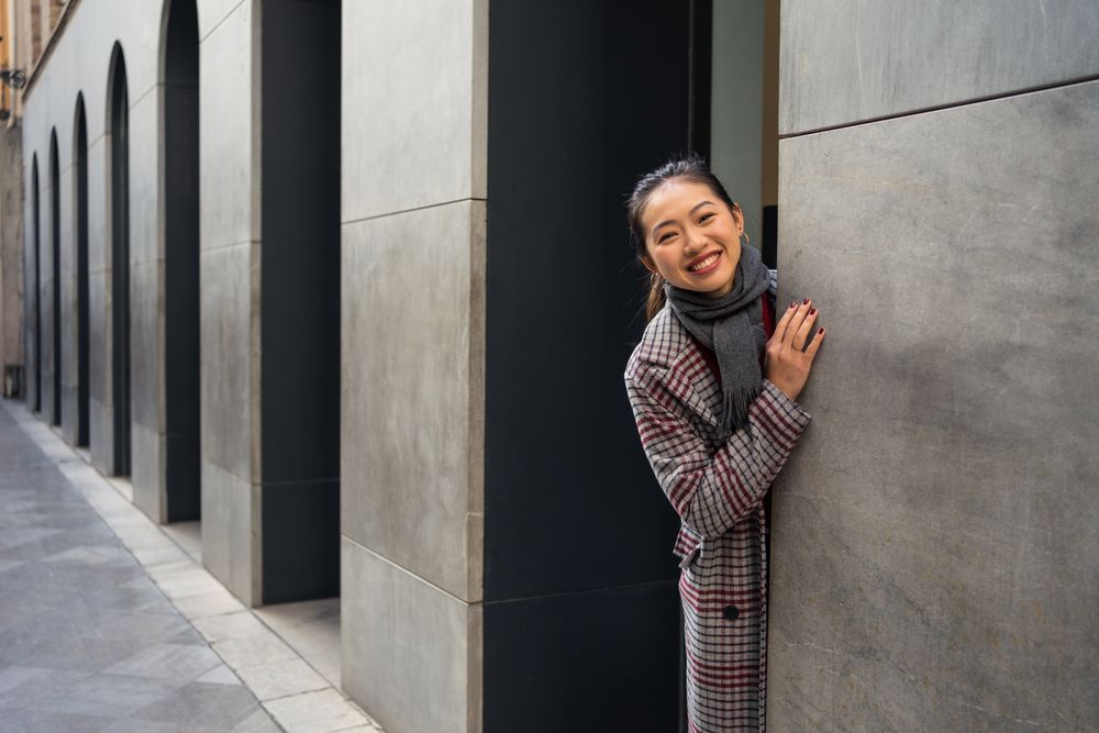 Joyful Asian resting female in casual clothing looking out modern wall of building at city street