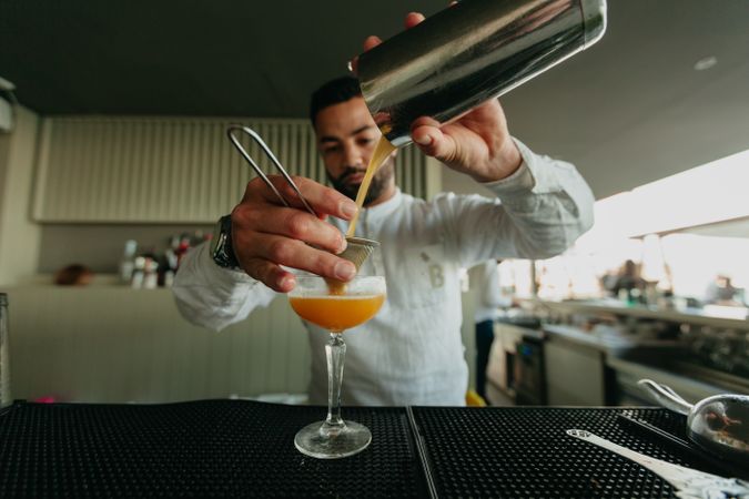 Barman pouring cocktail and alcoholic drinks from shaker in a bar.