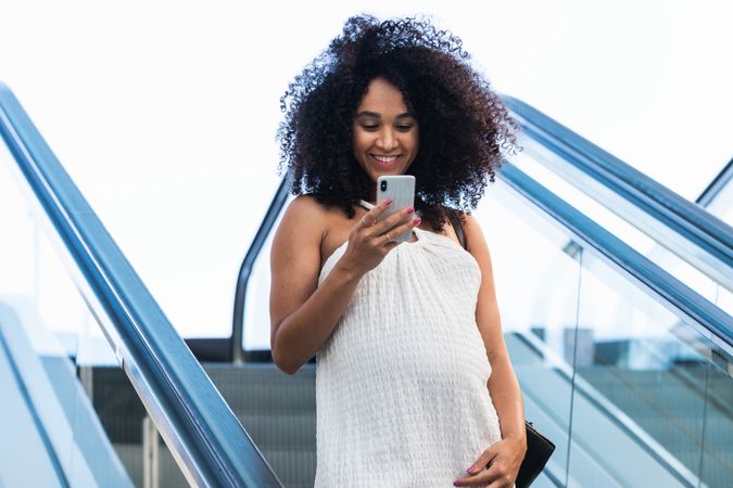 Pregnant Black woman browsing smartphone on escalator