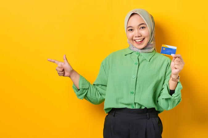 Smiling Muslim woman in headscarf and green blouse holding credit card and pointing aside
