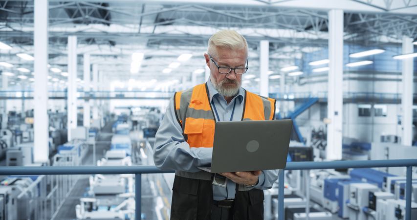 Engineer Standing on a Platform, Using Laptop Computer at an Electronics Factory. Machines are Undergoing Maintenance, Specialist Monitoring the Updates Through Online Software
