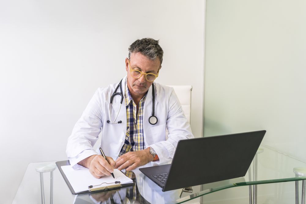 Male doctor working on laptop and clipboard in medical office