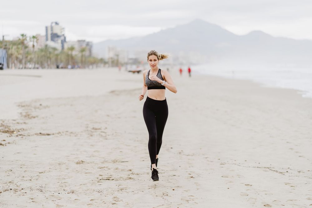 Female runner in stylish sportswear jogging along calm sandy seaside