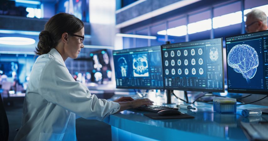 White Female Neuroscientist Using Desktop Computer To Analyze CT Scans Of Human Brain In Busy Research Center Laboratory.