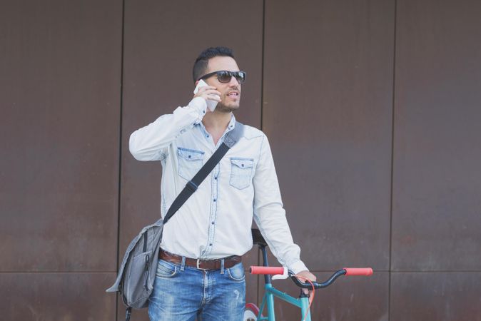 Male in sunglasses standing with red and green bicycle and looking around speaking on phone