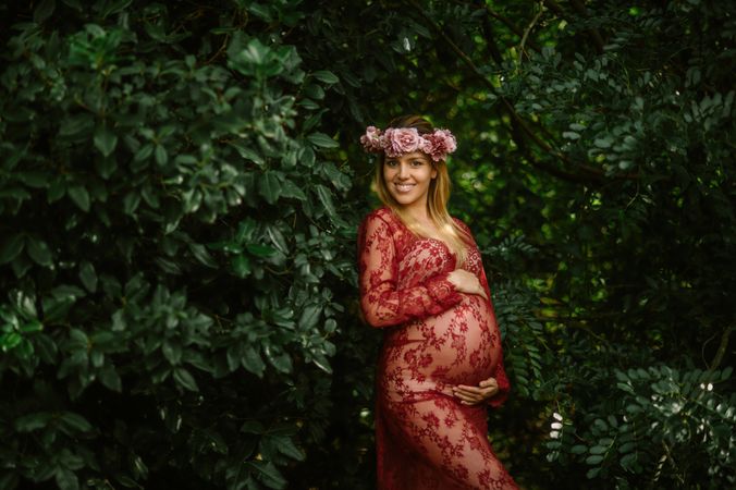 Pregnant woman with closed eyes touching belly and chin while standing in garden in sunny day