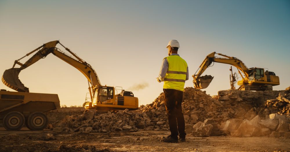 Male Urban Planner Wearing Protective Goggles And Using Tablet On Construction Site On A Sunny Day. Man Inspecting Building Progress. Excavator Loading Materials Into Industrial Truck
