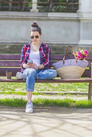 Front view of a young hipster woman sitting on a park bench relaxing in a sunny day while looking away