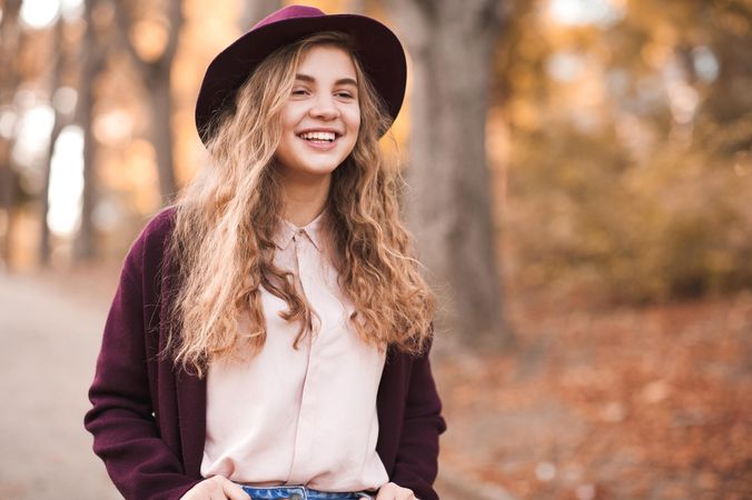 Happy teenage girl in purple coat with hat standing near fallen autumn tree leaves