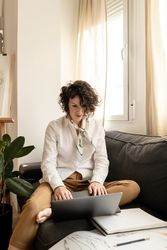Woman using laptop near draws on papers on sofa in room