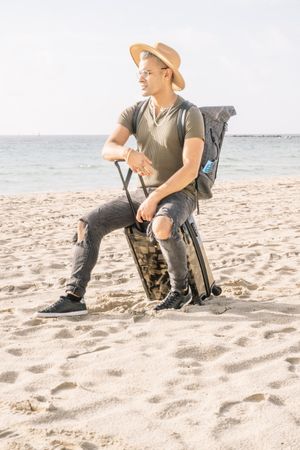 young latino man with a modern look and hat on miami beach with a travel suitcase
