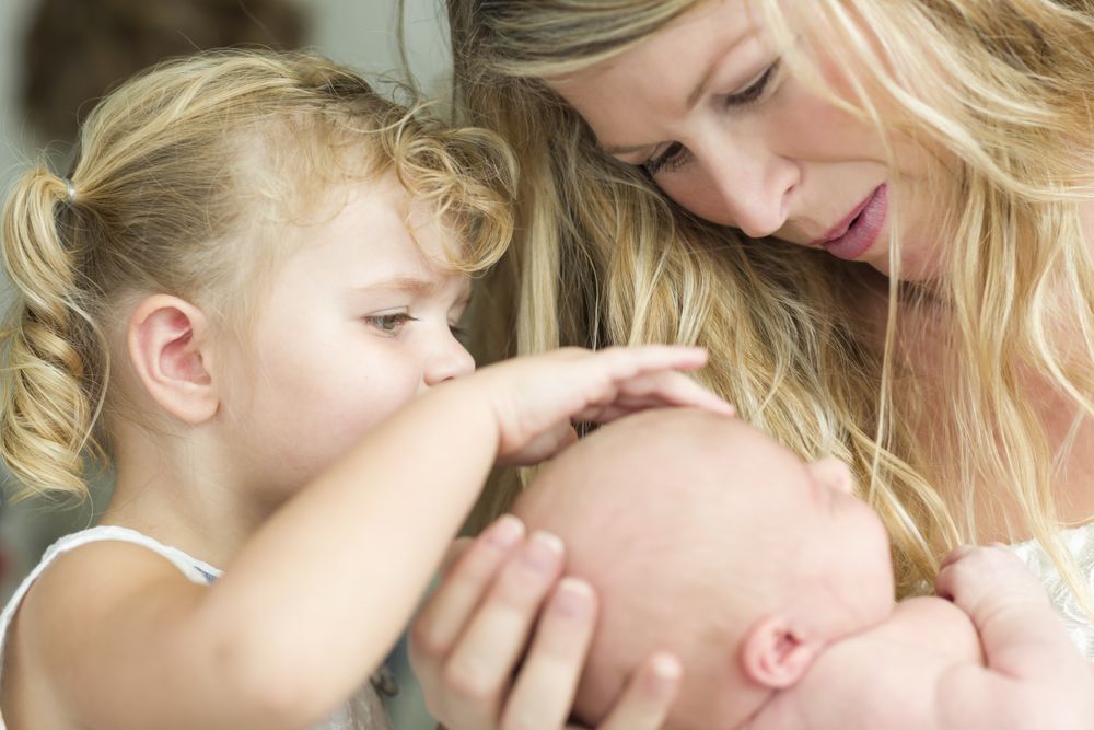 Attentive Mother Holds Newborn Baby Girl as Young Sister Looks