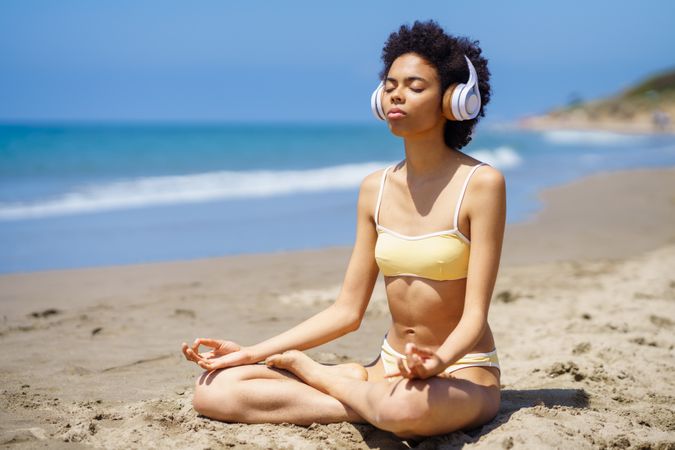 Woman in yellow swimwear sitting cross legged and meditating on beach in head phones
