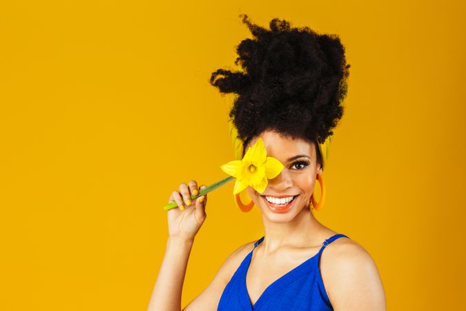 Portrait of happy Black woman with large earrings holding a daffodil over her eye