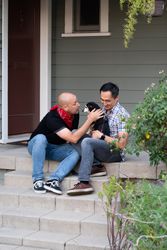 Married couple sit with their dog on front porch of their home with plants in foreground