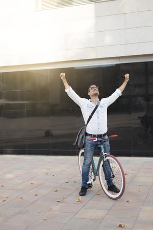 Joyful male sitting on colorful bicycle with both arms up and outstretched