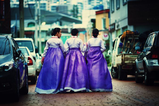 Back view of three Korean women in hanboks walking side by side between cars