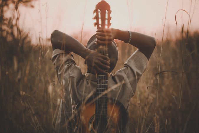 Man holding classical guitar while sitting on grass field during sunset