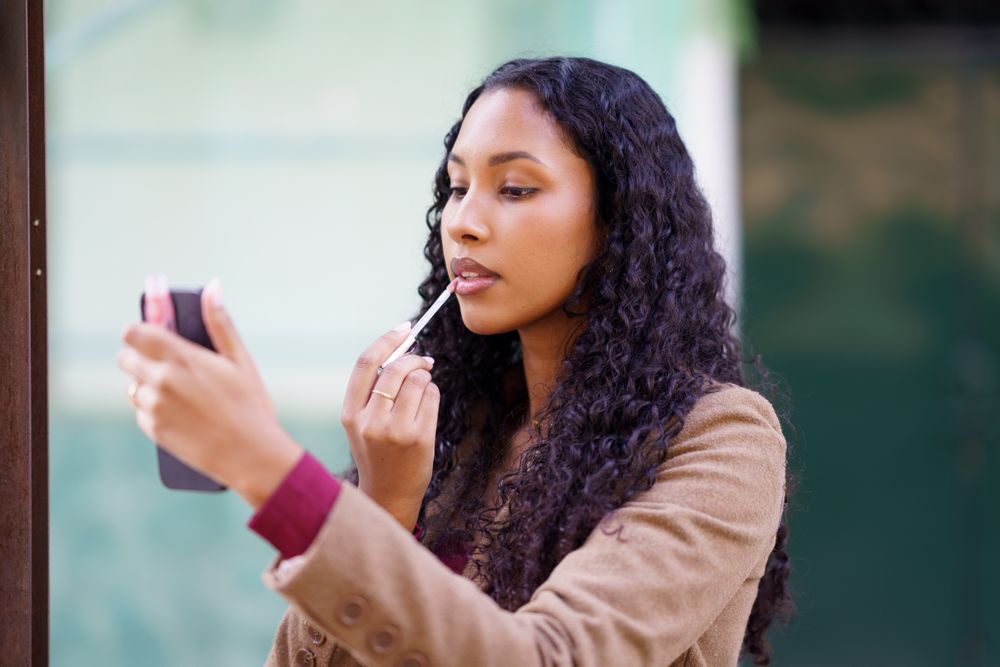 A woman is applying lip gloss while looking at her smartphones mirror, showcasing her beauty
