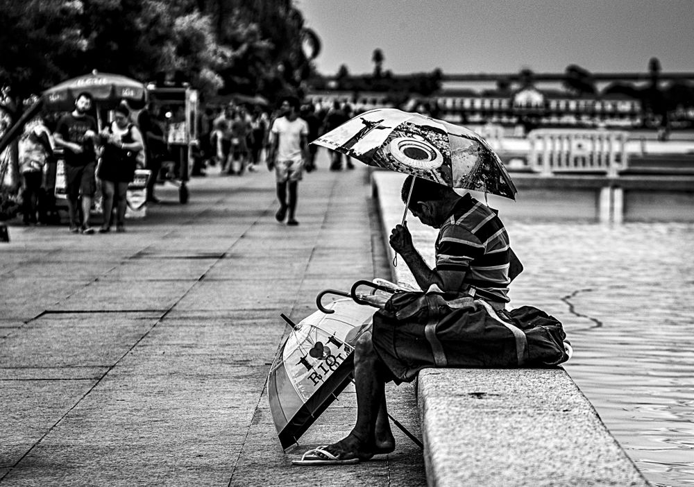 Grayscale photo of a man holding an umbrella and sitting on concrete bench
