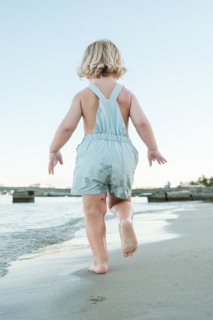 Little barefoot toddler in summer suit walking on sandy beach