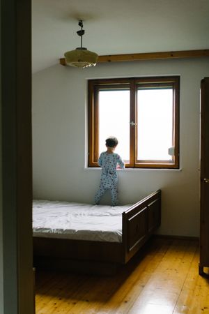 Child in pajamas standing on bed and looking out window