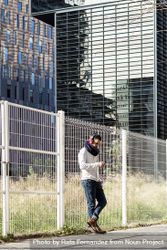 Male in headphones holding smartphone while leaning on a metallic fence outside 4d8adD