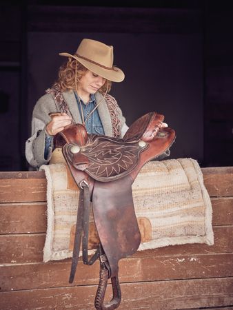 Woman in cowgirl hat placing saddle on carpet in stall