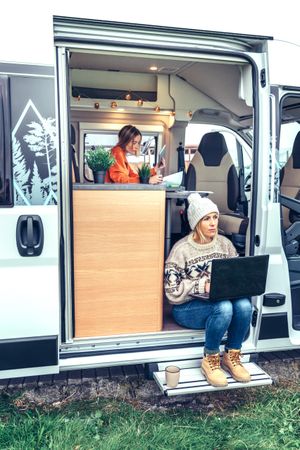 Female in sweater working remotely sitting on step of van while on a road trip with friend, vertical