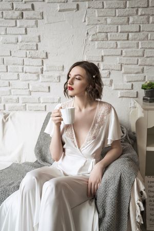 Pensive adult woman looking away and dreaming while sitting on sofa and having hot drink in light modern living room
