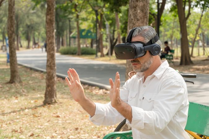 Mature grey haired man sitting outside in park with hands up wearing VR headset