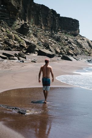 Back of man in swim trunks and cap walking along beach